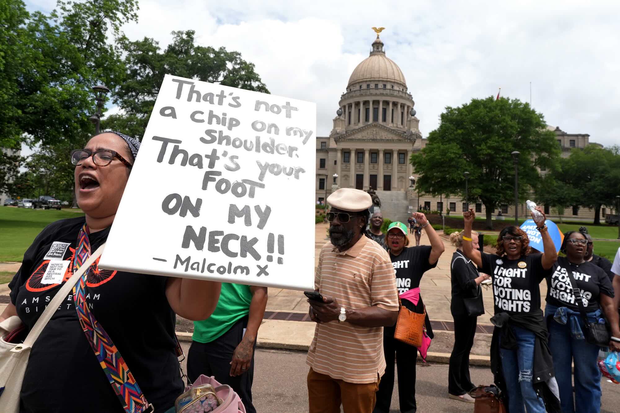 People march past the Mississippi capital building, some in shirts that read 'I Fight For Voting Rights'. A person holds a sign that reads 'That's not a chip on my shoulder, that's your foot on my neck!! - Malcom X'