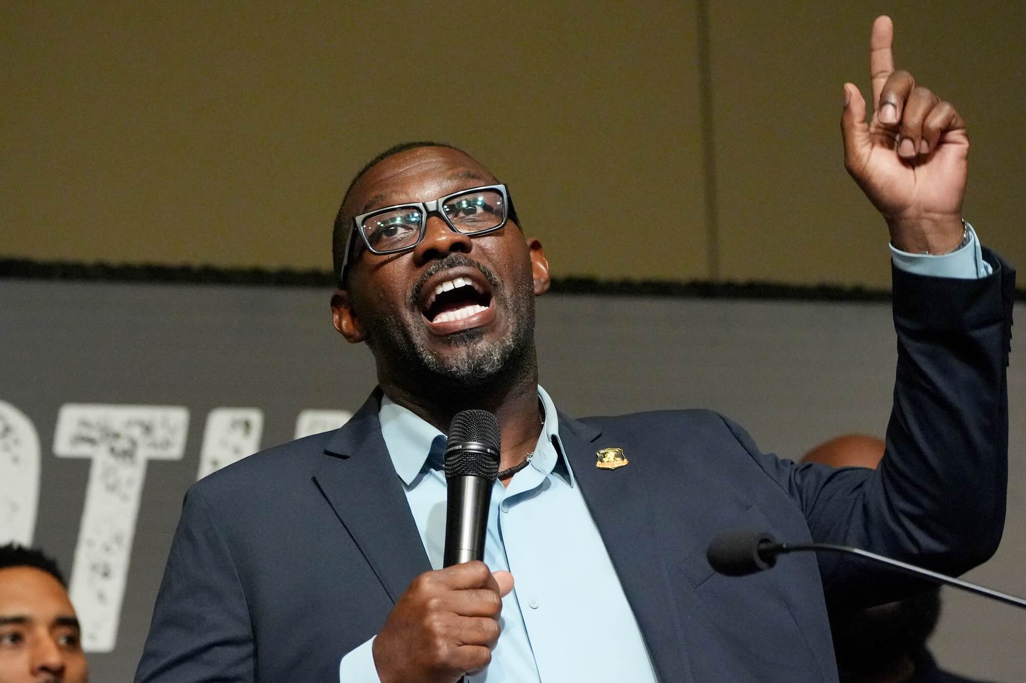 A closeup of Bryant Clark speaking on stage, mic in one hand and the other hand pointing up