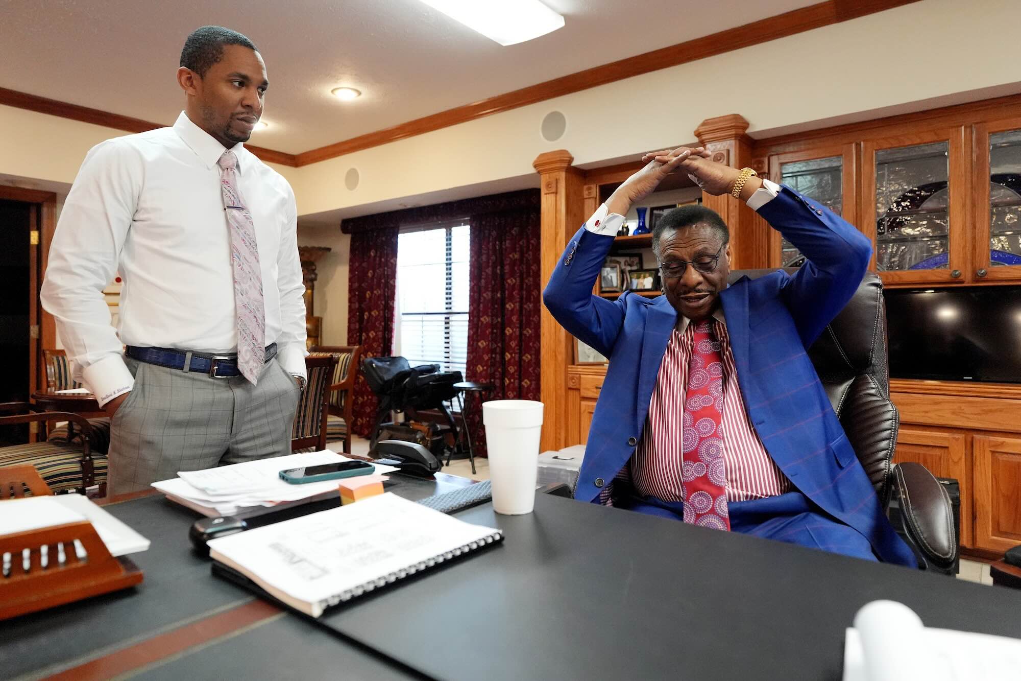 Edward Blackmon sits at his desk, arms stretched above his head, while Bradford Blackmon stands beside the desk looking at him