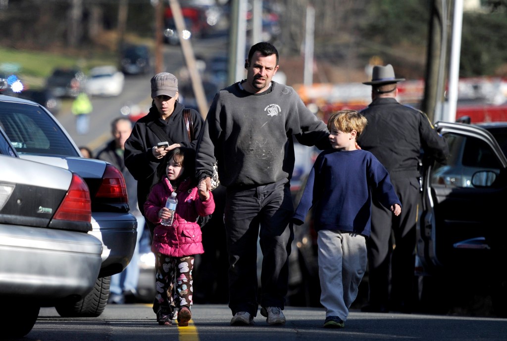 A woman and man walking with two children on a street with several parked cars.