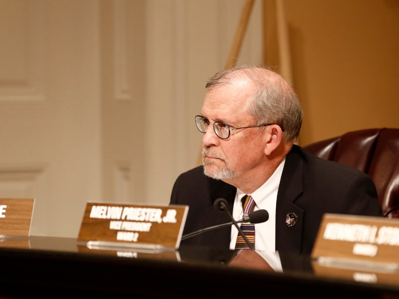 An older man wearing glasses and a suit sits inside the city council room