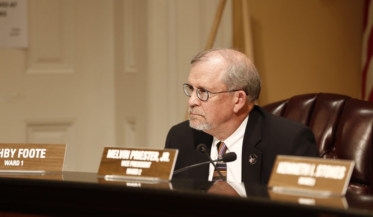 An older man wearing glasses and a suit sits inside the city council room