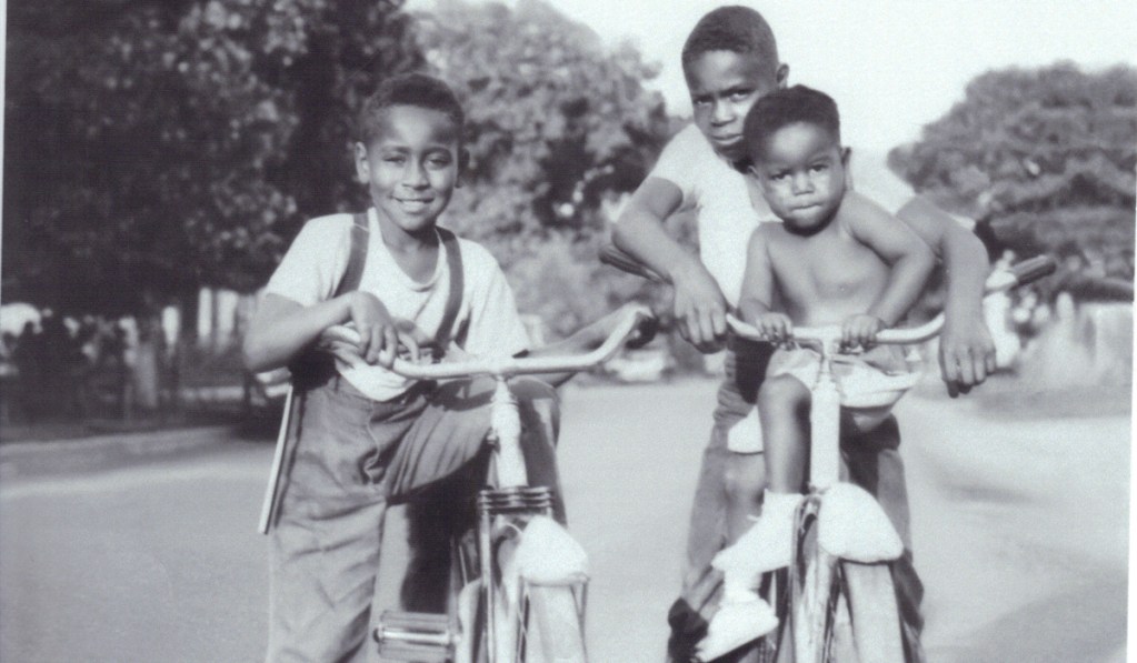 Two boys who look like they're around 10 ride bicycles in an old black and white photo, one with a much younger boy sitting up front holding the handle bars