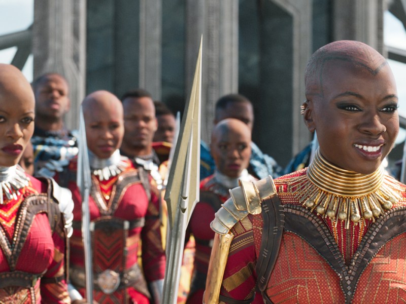 A still from a movie depicting many bald women with red outfis and metal worked details, with matching metal spears