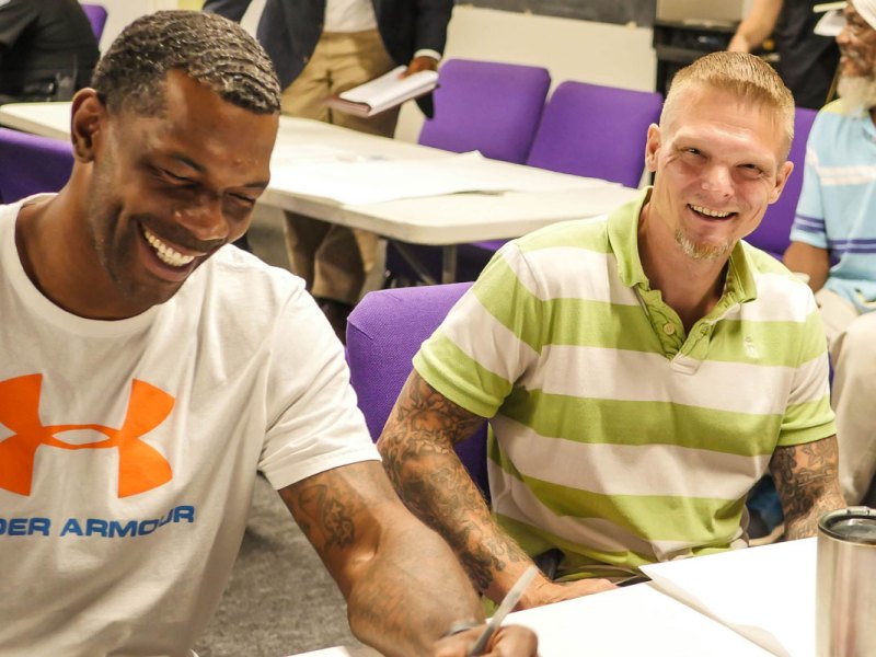 a photo of a black man and a white man smiling and laughing while sitting at a table together