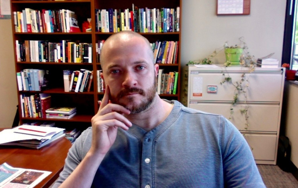 James Thomas sitting behind an office desk, shelves visible behind him