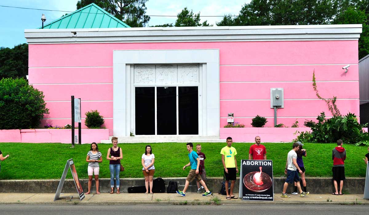 People stand in a line outside a pink building.