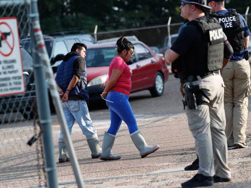 Men in Police/ICE vests watch as two factory workers i work boots talk with their wrists ziptied