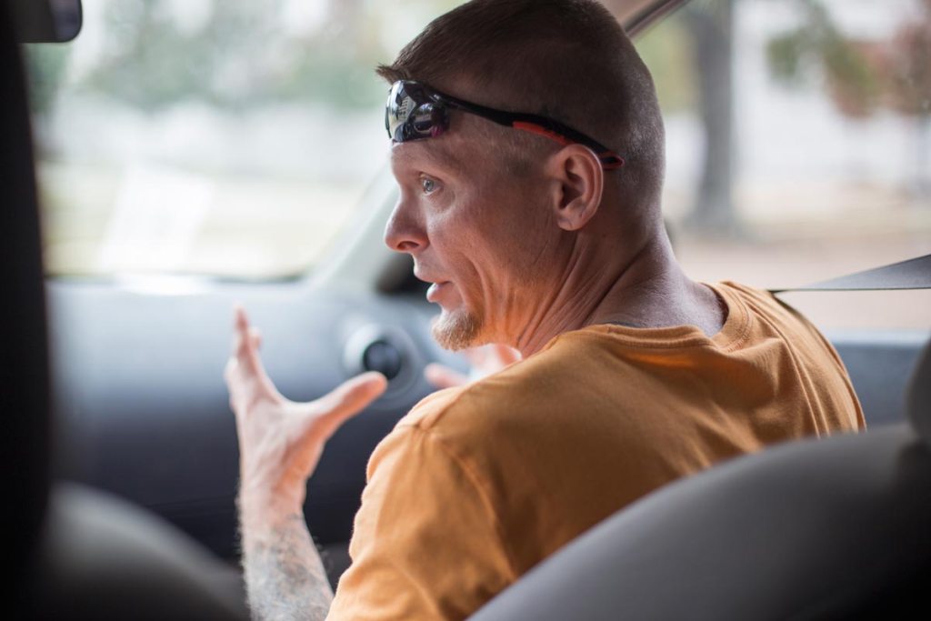 A white man in an orange shirt speaking in the passenger seat of a car.