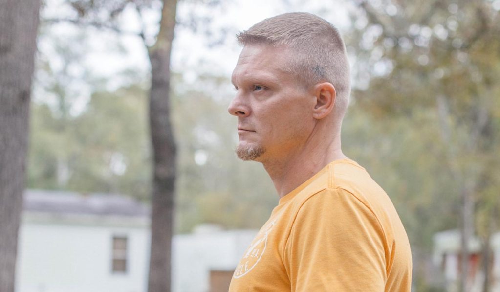 a photo shows a white man with short hair in a yellow shirt standing outside with trees behind him