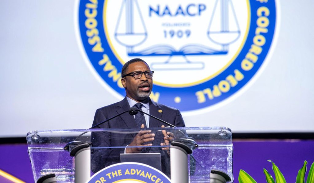 A man in a suit at a clear podium in on a white and blue stage covered in the NAACP logo