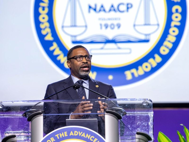A man in a suit at a clear podium in on a white and blue stage covered in the NAACP logo