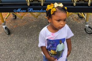 Leah Davis, a small Black child with yellow ribbons in her hair, stands in front of her father’s casket in June 2017. She is wearing a white graphic t-shirt with a photograph of her father in the center with pink candy stains around her collar.