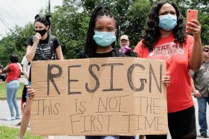 A young Black girl, wearing a mask, holds a cardboard sign as she stands outside Petal City Hall protesting Mayor Hal Marx for his comments. The sign reads: "Resign: This is Not the First Time"