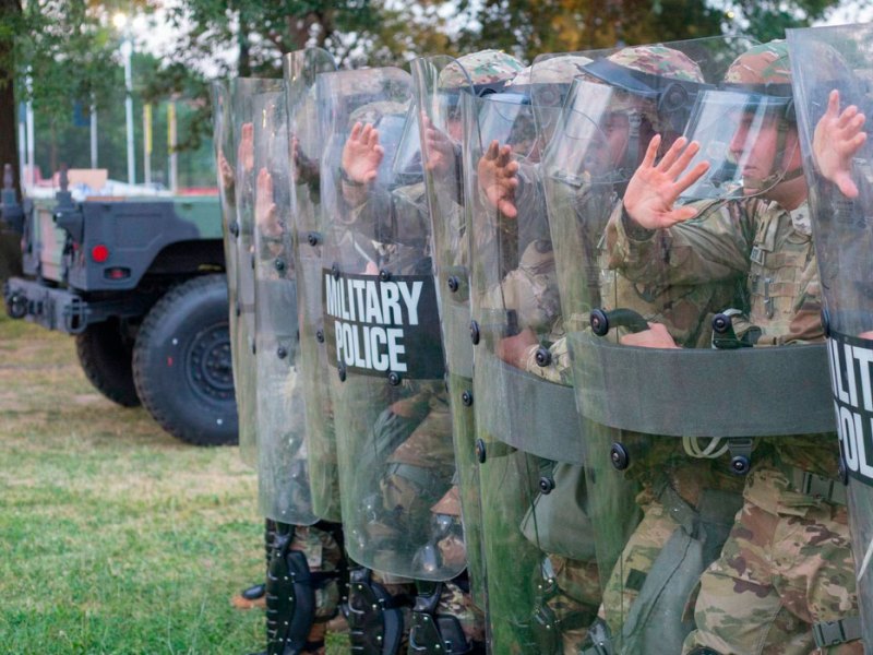 A row of men in camouflage uniforms hold up shields that read 'Military Police'
