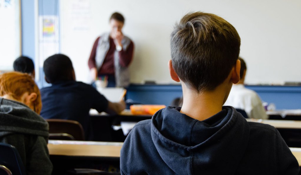 A young student looking forward to a man teaching at the front of a classroom.