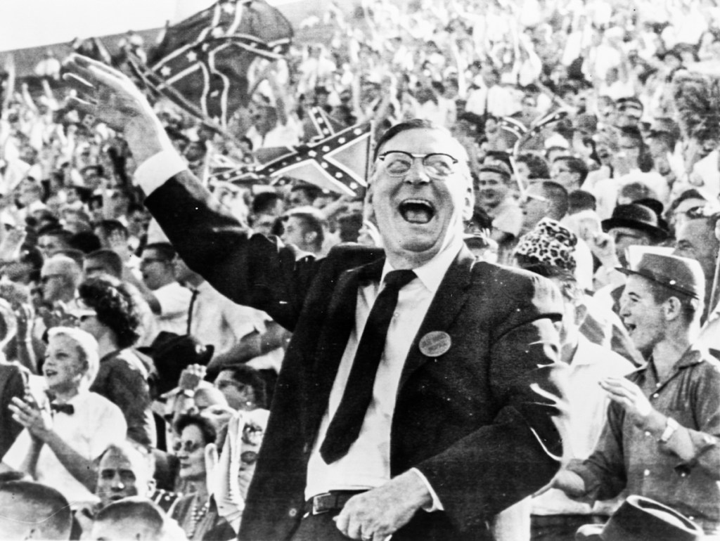 A black and white photo of a man at a football game as the crowd cheers and waves Confederate flags