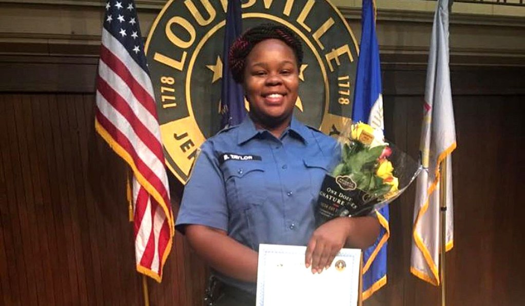 A woman in a uniform stands in front of a Louisville sign holding flowers and a certificate.