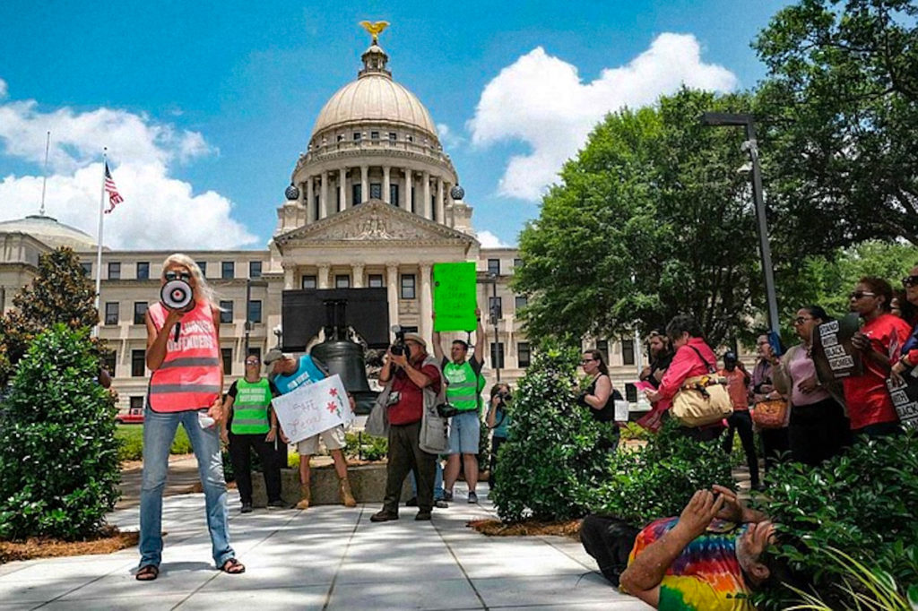 a photo shows a woman with a megaphone wearing a vest that says "Pink House Defender" while other people holding signs stand behind her in front of the Mississippi Capitol Building