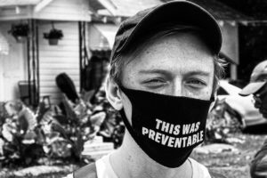A white man wears a mask with the words: "This was preventable"