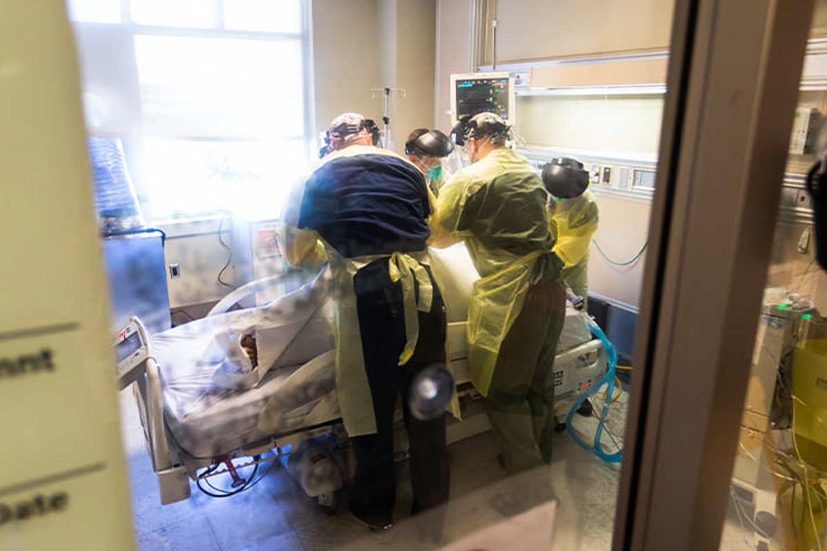 Four healthcare professionals in full PPE surround a hospital bed as they work on a COVID-19 patient.