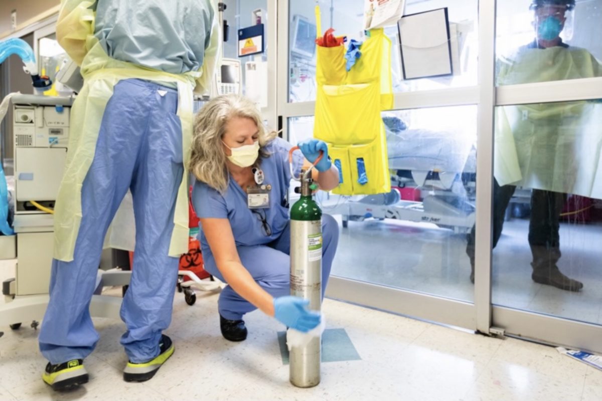A blonde female respiratory therapist crouches down to prepare an oxygen tank for use by a hospitalized COVID-19 patient.