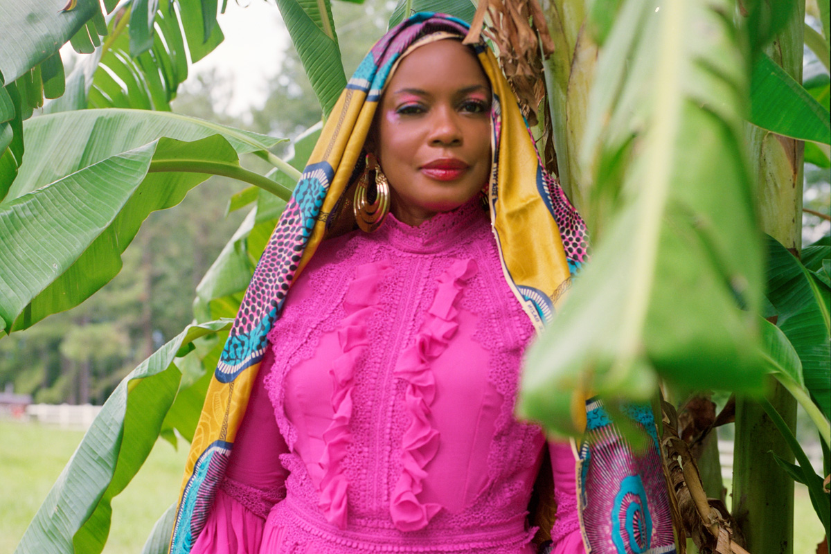 Aunjanue Ellis poses outside under a banana plant tree wearing a pink dress and a bold African patterned shawl