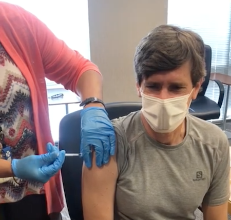 Dr. Dobbs, wearing a mask, is injected with the COVID-19 vaccine by a healthcare professional wearing blue gloves.