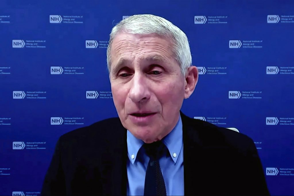 Dr Anthony Fauci in blue button down shirt and black jacket, talking into the camera. A blue background with repeating NIH logos is behind him