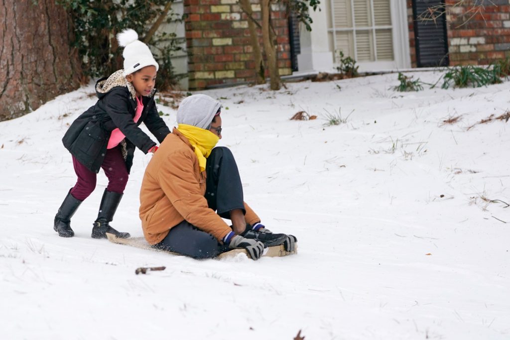 Two children in a snowy front yard, sledding on a cardboard box