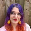Kristin is seen with multicolored hair, large yellow lemon earrings, and a rainbow striped sweater