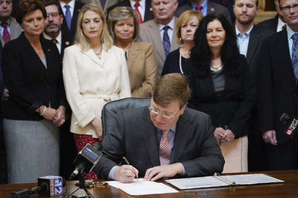 a photo of Tate Reeves signing a bill with lawmakers standing behind him
