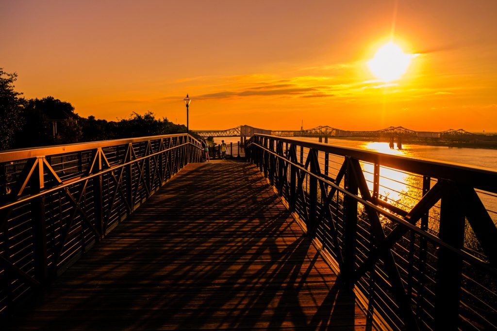 A walkway in Natchez, Mississippi is covered in shadows as the orange sets over the Mississippi River in an orange sky