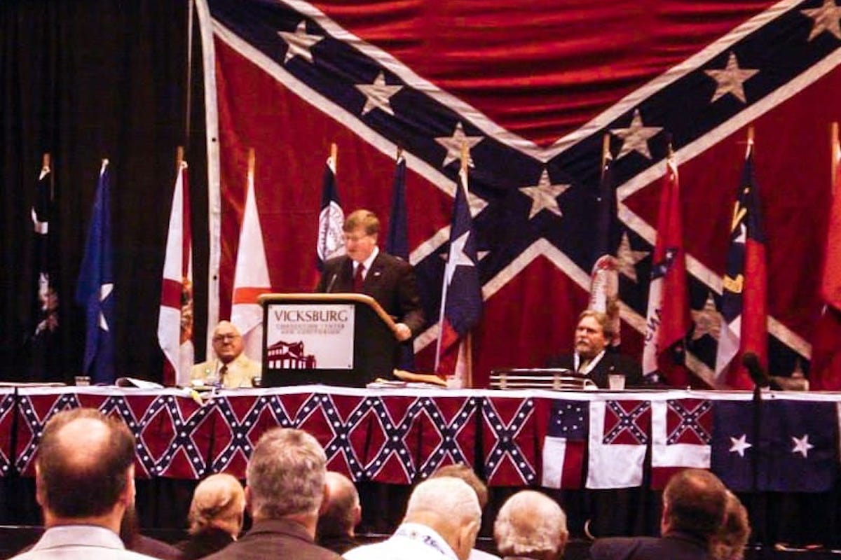 Tate Reeves speeking at a podium surrounded by Confederate flags.
