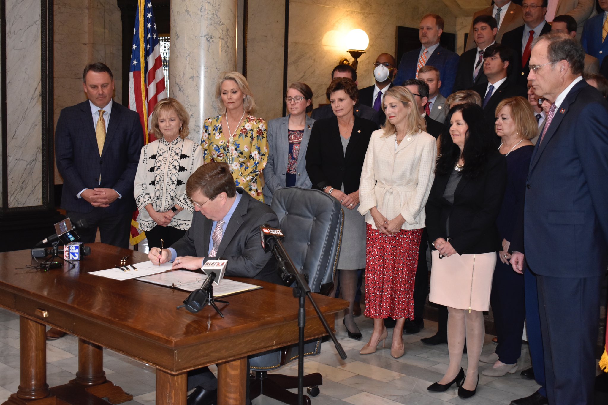 Governor Tate Reeves is seen sitting at a desk signing a ban on transgender athletes into law on March 11, 2021 with supporters of the legislation standing around him.