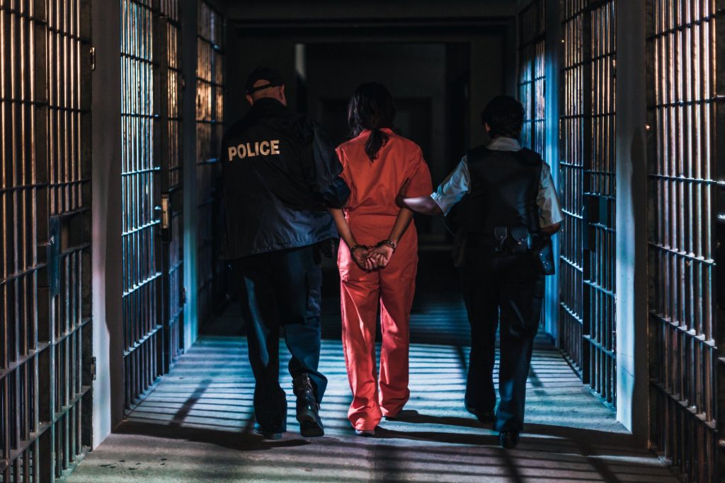 A photo shows a woman in an orange jump suit led through a corridor of cells by two guards
