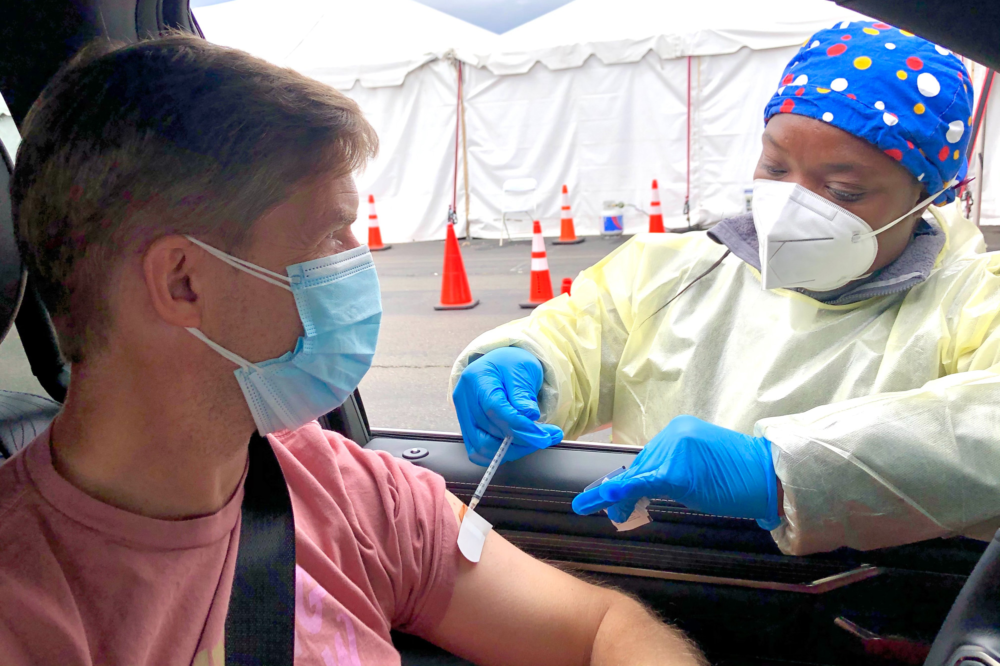 A young, Black female healthcare professional wearing PPE administers a COVID-19 vaccine injection to a middle-aged white male through his vehicle's window.