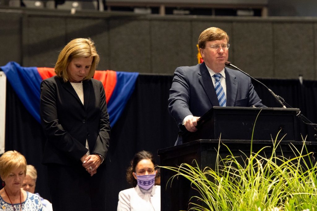 Governor Reeves stands at a podium in the Mississippi coliseum with wife Elee Reeves next to him and the words "National Day of Prayer" behind him