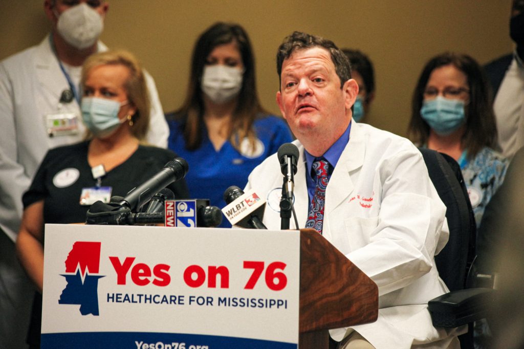 Dr. John Gaudet stands at the podium with nurses and health workers behind him and a YES ON 76 campaign sign in front of him in support of Medicaid expansion