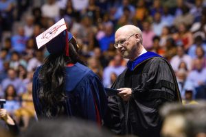 Will Norton, seen here in 2018 as the journalism school dean, hands a diploma to a student at a crowded graduation ceremony