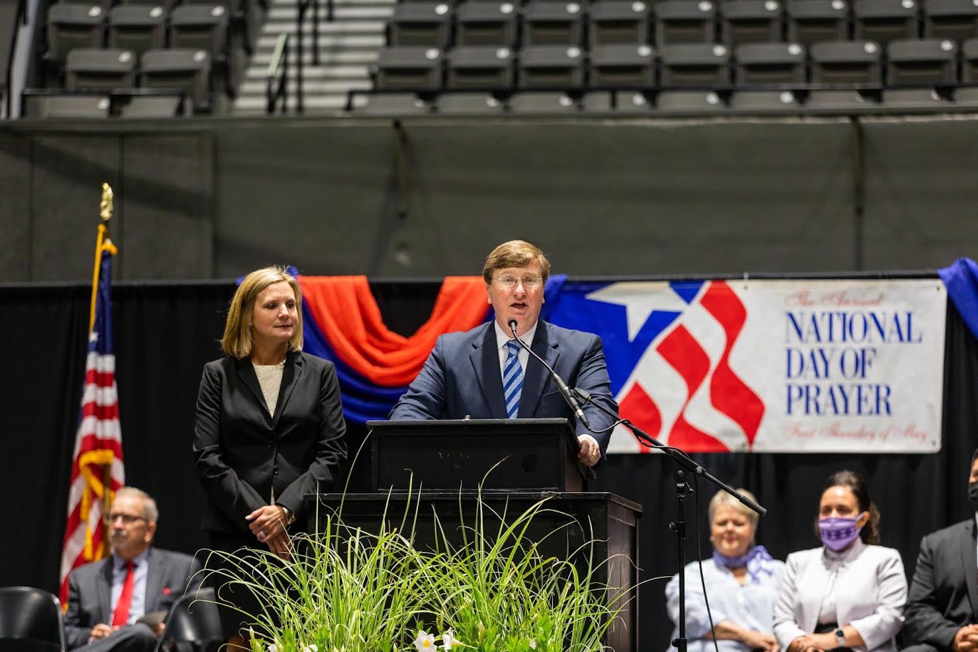 Governor Reeves stands at a podium in the Mississippi coliseum with wife Elee Reeves next to him and the words "National Day of Prayer" behind him