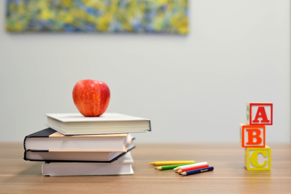 3 books stacked on a desk with a red apple on top, some color pencils and ABC blocks on a desk (teachers)
