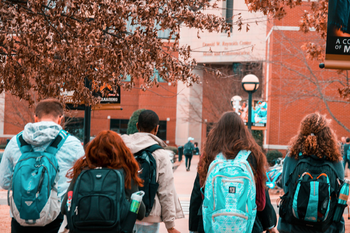 Kids with backpacks are seen from behind walking to the steps of a redbrick school building