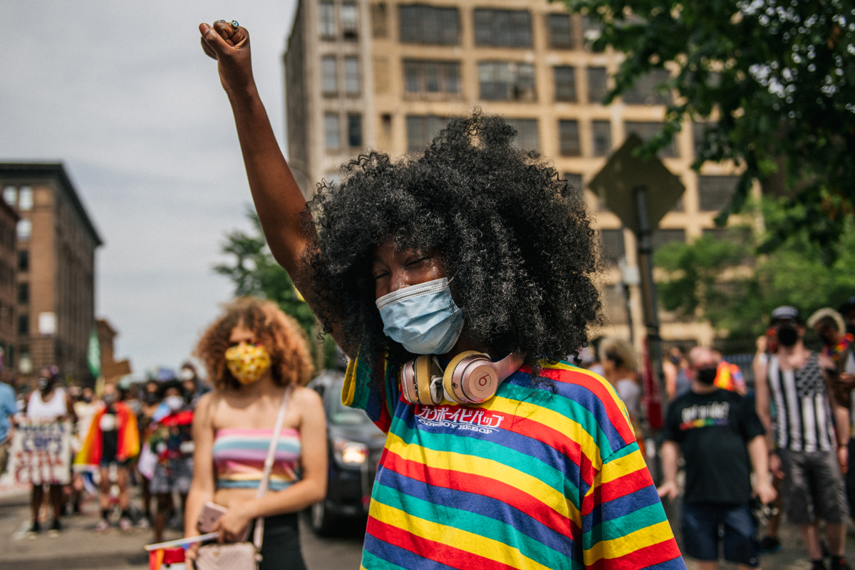 A young Black woman in a colorful striped t-shirt and medical mask with a fist raised in the foreground of a protest