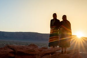 Two Indigenous women standing side by side on red stones in front of a sunset