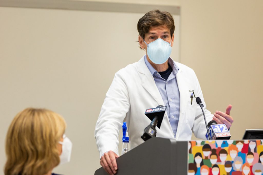 Dr. Thomas Dobbs, masked and wearing a lab coat, speaking at a podium