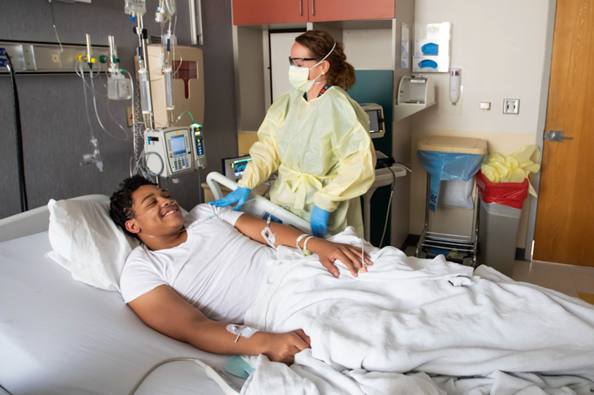 A 16 year old boy lies in a hospital bed while a nurse checks on him