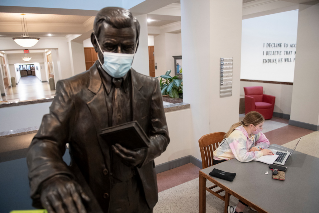 a student wearing a masks studies in the library next to a masked statue of a man