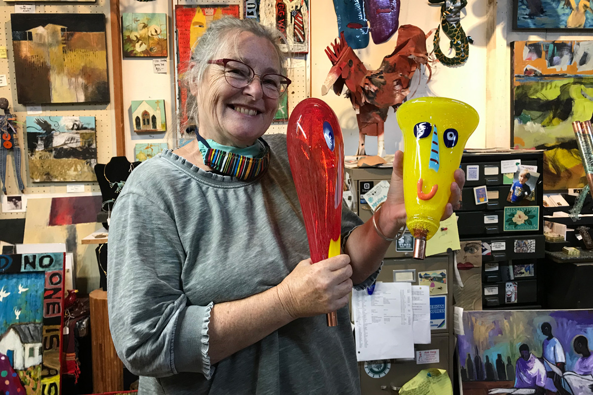 Ginger Kelly in grey holding two pieces of glass artwork. The pieces are red and yellow and have faces.
