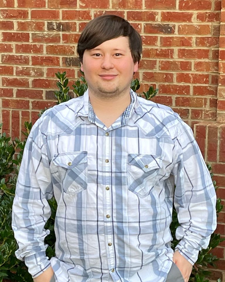 Nate Schumann in a long sleeve white and blue plaid shirt standing in front of a brick wall
